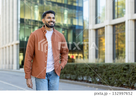 Young bearded indian businessman walking confidently near modern office buildings, smiling while looking away, enjoying the vibrant atmosphere of a successful career and future aspirations 132997652