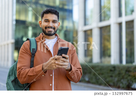 Young latin university student smiling while using a smartphone app near a campus building, dressed in casual clothes and carrying a green backpack, enjoying a vibrant day outdoors Young latin university student smiling while using a smartphone app near a campus building, dressed in casual clothes and carrying a green backpack, enjoying a vibrant day outdoors 132997675