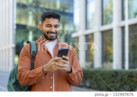 A happy young adult male browsing smartphone while wearing a backpack outdoors in the urban environment. The sunlight highlights his cheerful expression, suggesting communication, study, or travel 132997676
