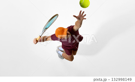 Focused tennis player in burgundy clothing jumping for serve on minimal white background 132998149