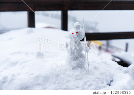 small snowman stands on a snowy surface and is holding a stick. The background is blurred with a winter landscape, winter fun small snowman stands on a snowy surface and is holding a stick. The background is blurred with a winter landscape, winter fun 132998550