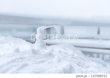 small snowman stands on a snowy surface, his nose an icicle. The background is blurred by a wintry atmosphere. Snow covers the ground, creating a serene winter scene, copy space 132998552