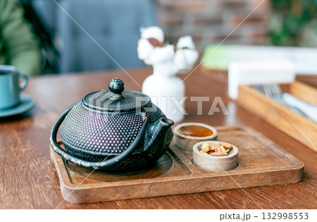 heavy black cast-iron teapot sits on a wooden tray. On it are two small bowls of honey and nuts. In the background is a white vase of cotton flowers 132998553
