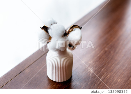 A white ceramic vase holds cotton branches with fluffy white bolls. The vase is placed on a wooden surface, creating a minimalist decor scene. A white ceramic vase holds cotton branches with fluffy white bolls. The vase is placed on a wooden surface, creating a minimalist decor scene. 132998555