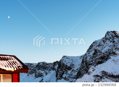 Snow-covered mountains under a clear blue sky with a small red house in the foreground. The moon is visible in the sky, adding to the serene winter landscape, copy space Snow-covered mountains under a clear blue sky with a small red house in the foreground. The moon is visible in the sky, adding to the serene winter landscape, copy space 132998568
