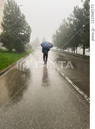 person walks alone on rainy street, holding blue umbrella. Trees line the road, and the ground is wet from the rain. Fog obscures the background, autumn person walks alone on rainy street, holding blue umbrella. Trees line the road, and the ground is wet from the rain. Fog obscures the background, autumn 132998573