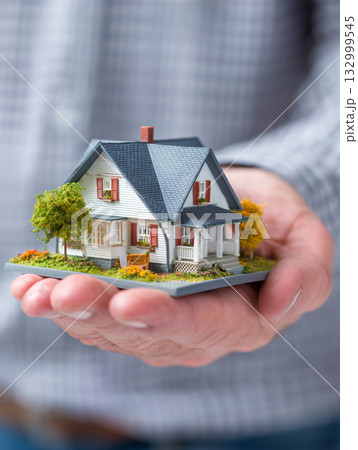 A close-up shows a man hand gently holding a small, detailed model house, against a soft, blurred background.   132999545