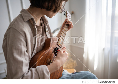 Young woman rehearsing ukulele chords at home, refining finger placement and strumming technique Young woman rehearsing ukulele chords at home, refining finger placement and strumming technique 133000646