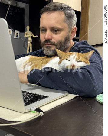 Bearded man working on laptop at home with ginger and white cat sleeping on computer desk, man hugging cat and looking at screen 133000909