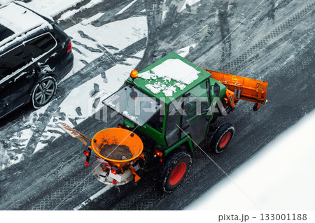Above view small green tractor orange snow plow salt spreader clears icy street during winter in city. Fresh tire marks footprints on gray asphalt contrast with white snow and bright machine colors. 133001188