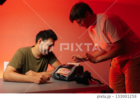 Two cheerful male content creators sitting at workspace, counting earnings near professional cameras under red light, showcasing successful digital entrepreneurship, studio shot, isolated background Two cheerful male content creators sitting at workspace, counting earnings near professional cameras under red light, showcasing successful digital entrepreneurship, studio shot, isolated background 133001258