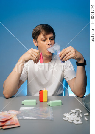 Vertical portrait of drug dealer smoking heated tobacco from sleek modern heat-not-burn tobacco device sitting at desk with money, pills and various drugs, exhaling smoke on blue background. Vertical portrait of drug dealer smoking heated tobacco from sleek modern heat-not-burn tobacco device sitting at desk with money, pills and various drugs, exhaling smoke on blue background. 133001274