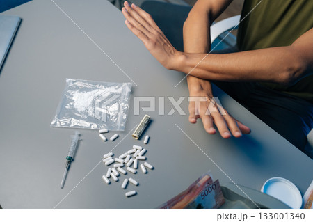 Recovering drug user pushing away narcotics, surrounded by scattered medical tools and paraphernalia, representing personal battle against substance dependency. Concept of healthy lifestyle. Recovering drug user pushing away narcotics, surrounded by scattered medical tools and paraphernalia, representing personal battle against substance dependency. Concept of healthy lifestyle. 133001340