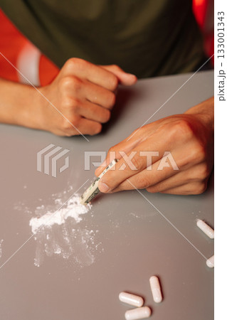 Vertical cropped shot of male hands preparing dose of cocaine with rolled banknote on gray table with scattered pills, illustrating dangers of drug abuse and addiction. Concept of substance abuse. 133001343