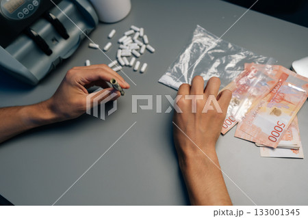 Drug dealer counting cash and preparing drug doses at night, surrounded by pills, money counter and laptop on table, highlighting dark world of illegal trafficking, close-up. 133001345