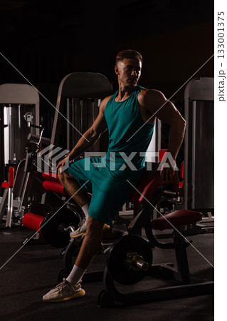 athlete showcases strength while sitting on gym equipment wearing workout gear. The setting is dimly lit with machines in the background highlighting focus and dedication to fitness. 133001375