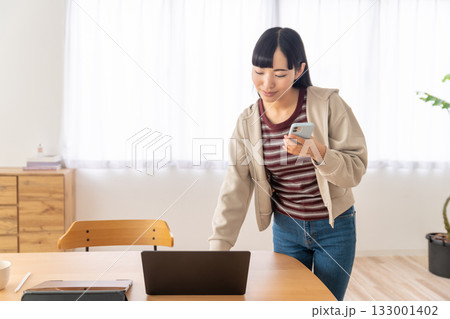スマホを片手に立ったままパソコンをチェックしている若い女性 スマホを片手に立ったままパソコンをチェックしている若い女性 133001402