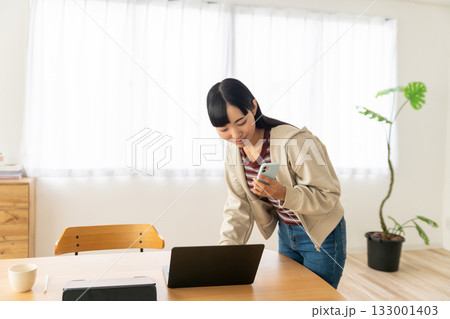 スマホを片手に立ったままパソコンをチェックしている若い女性 133001403
