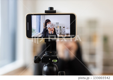 Caucasian woman in her thirties recording a financial vlog, dressed in a black business suit, holding dollar bills in hands, emphasizing wealth. Taken in a modern professional environment. 133003542