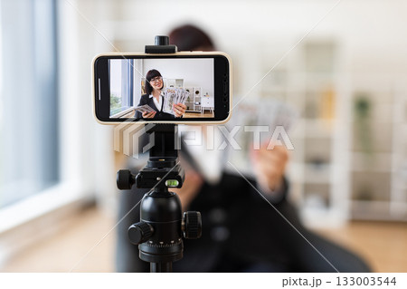 Caucasian young female professional blogging about financial tips, showing US dollar cash, seated in modern office with camera setup, wearing formal business attire, focusing on business communication 133003544
