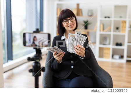 Caucasian young female professional blogging about financial tips, showing US dollar cash, seated in modern office with camera setup, wearing formal business attire, focusing on business communication Caucasian young female professional blogging about financial tips, showing US dollar cash, seated in modern office with camera setup, wearing formal business attire, focusing on business communication 133003545