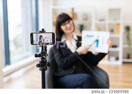 Caucasian woman in business suit recording video about revenue growth using graphs, modern office setup with natural light, professional presentation, concept communication and education. 133003560