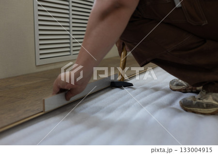 Man installing click-lock laminate flooring over white foam underlay, carefully tapping plank into place with hammer and tapping block during home renovation project. Man installing click-lock laminate flooring over white foam underlay, carefully tapping plank into place with hammer and tapping block during home renovation project. 133004915