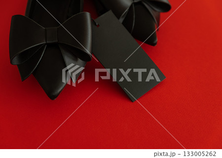 Elegant black pointed-toe shoes adorned with bows and a blank tag on red background Elegant black pointed-toe shoes adorned with bows and a blank tag on red background 133005262