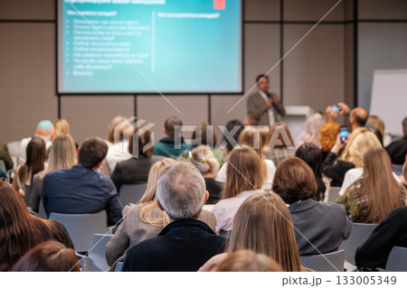 Engaged audience at a business conference listening to a speaker in a modern lecture hall with a large screen 133005349