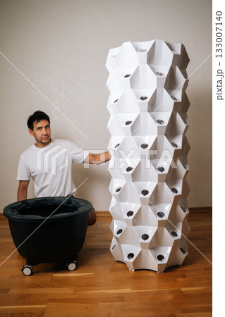 Vertical portrait of urban farmer assembling vertical hydroponic tower, growing fresh produce in compact indoor space with sustainable agricultural technology. Concept of home gardening. 133007140