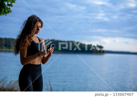 A Young Woman Enjoys the Beauty of Nature by the Tranquil Water While Using Her Smartphone 133007407