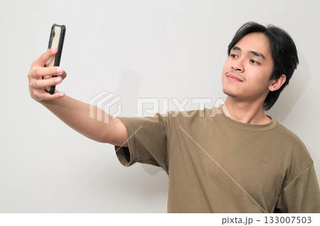 Portrait of handsome young Asian Thai man using phone taking selfie against white background 133007503
