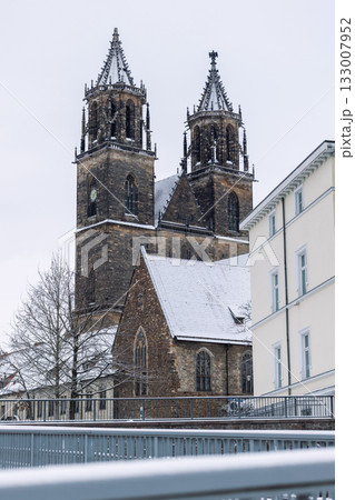 Snow-covered Magdeburg Cathedral towers rise against pale winter sky framed by modern building. Gothic architecture and frosty details create serene contrast of history and contemporary city scene Snow-covered Magdeburg Cathedral towers rise against pale winter sky framed by modern building. Gothic architecture and frosty details create serene contrast of history and contemporary city scene 133007952