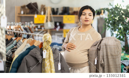 Pregnant mulatto young female client choosing trousers in clothing mall 133012381