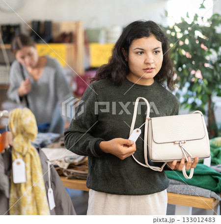 Latin girl is busy looking at clothes items in store, she chooses handbag 133012483