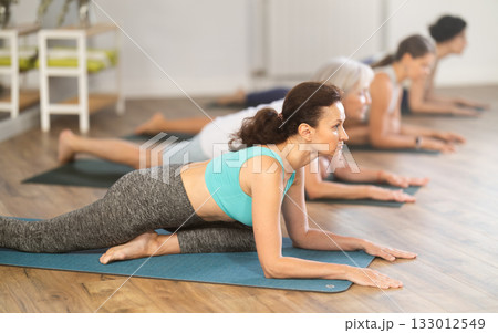 Group of women practicing pigeon pose of yoga on mat in fitness room Group of women practicing pigeon pose of yoga on mat in fitness room 133012549