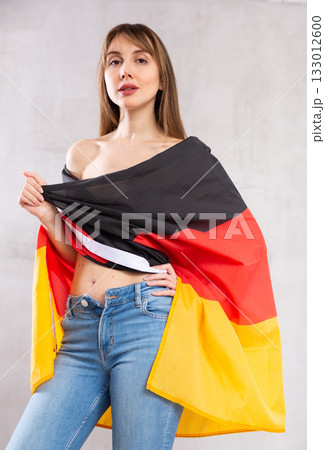 Flirting young woman in jeans with state flag of Germany against gray wall indoors 133012600