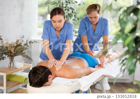 Two female masseuses perform stroking and rubbing of muscles of back and shoulders to guy client 133012818
