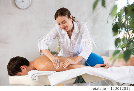 Professional young masseuse performing back massage for male patient Professional young masseuse performing back massage for male patient 133012835