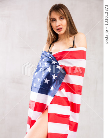 Serious patriotic young woman dressed in American flag against gray wall background indoors Serious patriotic young woman dressed in American flag against gray wall background indoors 133012873