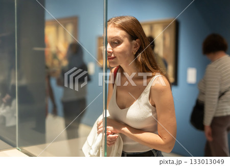 Young female visitor stands near glass display case and examines valuable exhibit 133013049