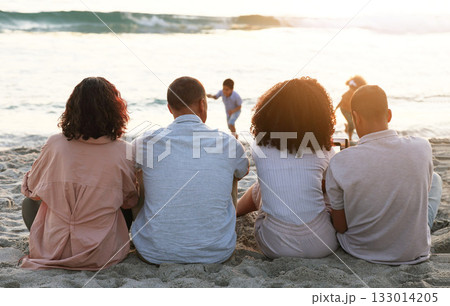 Family sitting on beach from the back, sunset and view of waves, zen and children playing in water together. Holiday, men and women relax in peace at evening ocean in Indonesia on tropical vacation. 133014205