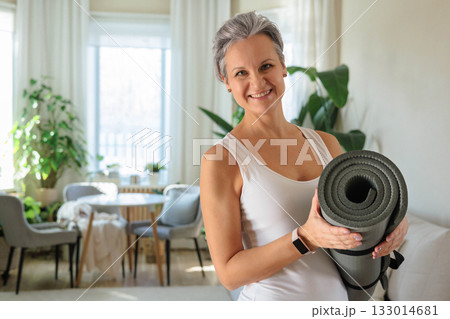 A beautiful smiling elderly woman with gray hair is holding a rolled-up yoga mat in her hands 133014681