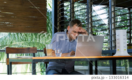 In a tropical setting a young man works on his laptop while sipping cold coffee and taking notes on the terrace  133016314