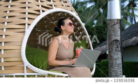 A young woman relaxes in a woven chair by the tropical beach working on her laptop and sipping a watermelon shake A young woman relaxes in a woven chair by the tropical beach working on her laptop and sipping a watermelon shake 133016338