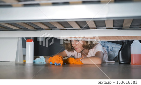 Girl in orange gloves lies down to clean under the bed at home 133016383