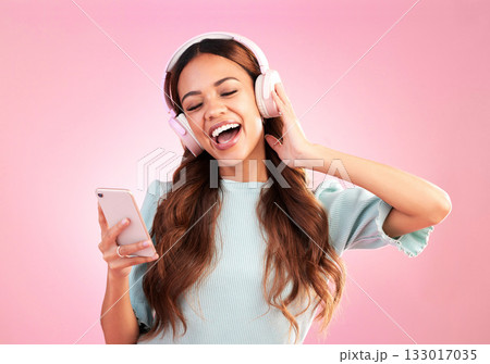 Music headphones, phone and woman singing in studio isolated on a pink background. Cellphone, radio singer and happy female with mobile streaming, enjoying and listening to audio, sound or podcast. 133017035