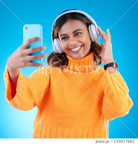 Music headphones, smile and selfie of woman in studio isolated on a blue background. Profile picture, radio and happy Indian female streaming, listening and enjoying sound, audio podcast or song. Music headphones, smile and selfie of woman in studio isolated on a blue background. Profile picture, radio and happy Indian female streaming, listening and enjoying sound, audio podcast or song. 133017663