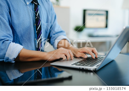 Young man typing on laptop and smartphone at modern office desk with coffee and tablet nearby. 133018574