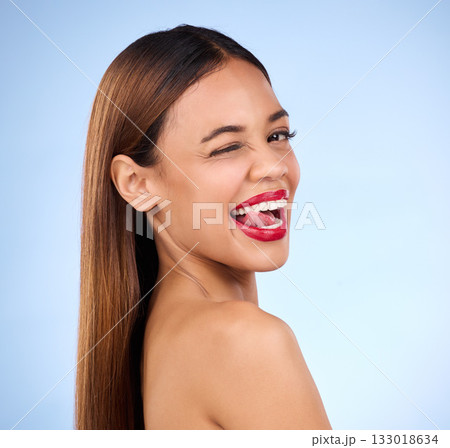 Beauty, woman and wink for face makeup and skincare cosmetics in studio. Female model person with tongue on teeth and red lips on a blue background for self care, facial glow and healthy skin shine 133018634